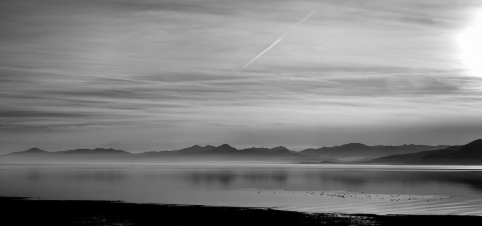 A black and white photo of a landscape. A cloudy sky stretches over mountains on the horizon, and in the foreground, a lake reflects the mountains and sky.